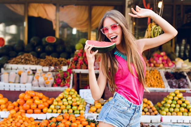 ragazza con i capelli lunghi sul mercato con il mercato dei frutti tropicali. Sta per assaggiare una fetta di anguria in mano