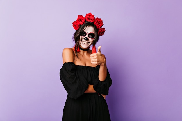 ragazza con il trucco del cranio che mostra il pollice in su. Donna con corona di rose in posa sulla parete viola