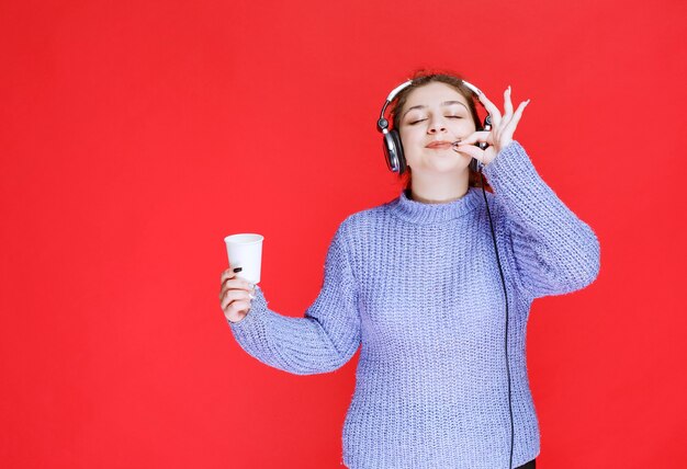 Ragazza con le cuffie che tiene una tazza di caffè e che mostra il segno di divertimento.