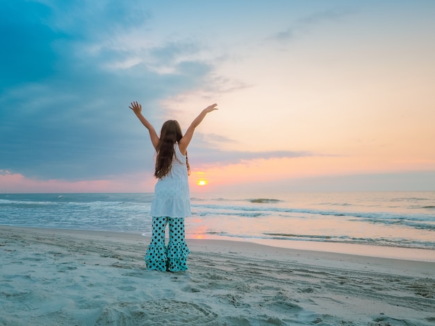 Ragazza con le mani alzate in piedi sulla spiaggia circondata dal mare durante il tramonto