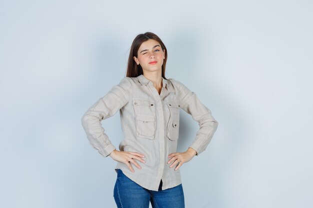 Ragazza con le mani sui fianchi, strizza l'occhio in camicia beige, jeans e sembra carina. vista frontale.