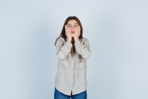 Ragazza con le mani sulle guance, tenendo la bocca aperta in camicia beige, jeans e guardando sorpreso, vista frontale.