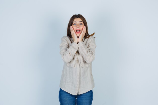 Ragazza con le mani vicino alla bocca, tenendo la bocca spalancata in camicia beige, jeans e guardando sorpreso, vista frontale.