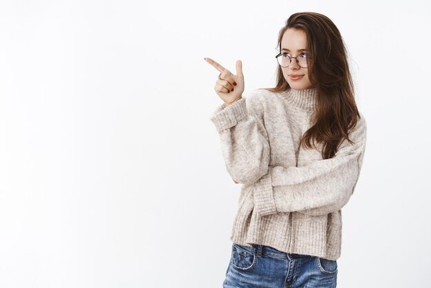 Ragazza con obiettivo puntato e mirato Ritratto di determinata donna bruna affascinante intelligente e attraente con occhiali e maglione che indica guardando a sinistra con curiosità e desiderio oltre il muro grigio