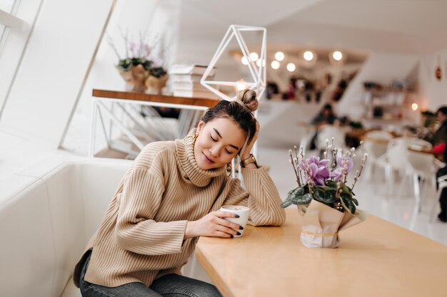 Ragazza con panino appoggiato su un tavolo di legno nella caffetteria Foto di donna in maglione leggero in posa con una tazza di caffè