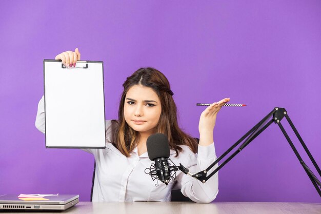 Ragazza confusa che tiene il suo taccuino in aria e guardando la fotocamera Bella ragazza seduta dietro la scrivania su sfondo viola Foto di alta qualità