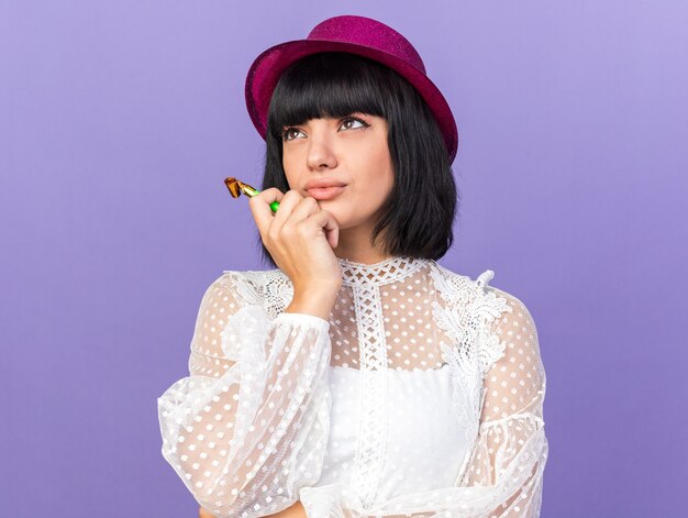 Ragazza confusa del partito che indossa il cappello del partito che tiene il corno del partito che cerca il mento commovente isolato sul muro viola