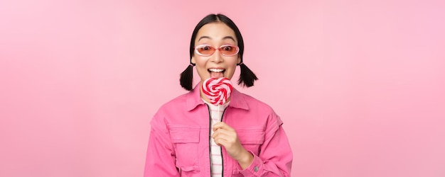 Ragazza coreana alla moda che lecca il lecca-lecca che mangia caramelle e che sorride in piedi in occhiali da sole su sfondo rosa