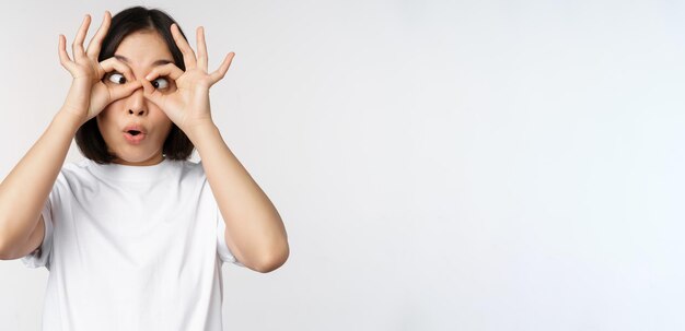 Ragazza coreana della giovane donna asiatica divertente che fa il gesto degli occhiali degli occhi che sembra felice alla macchina fotografica che si leva in piedi sopra il fondo bianco