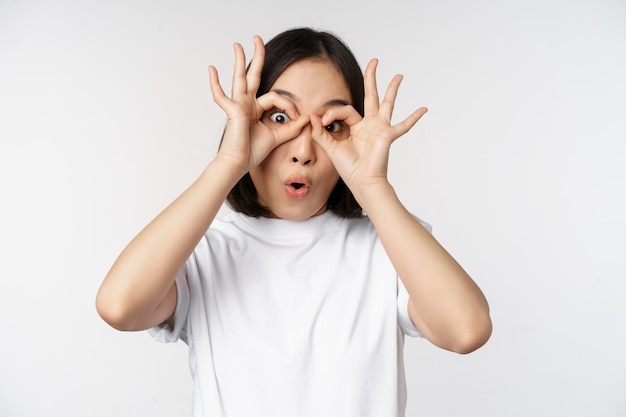 Ragazza coreana della giovane donna asiatica divertente che fa il gesto degli occhiali degli occhi che sembra felice alla macchina fotografica che sta sopra il fondo bianco