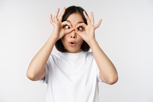 Ragazza coreana della giovane donna asiatica divertente che fa il gesto degli occhiali degli occhi che sembra felice alla macchina fotografica che sta sopra il fondo bianco