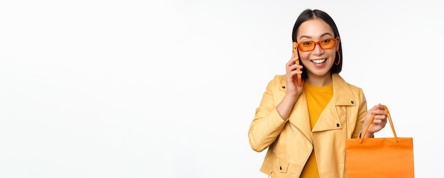 Ragazza coreana moderna sullo shopping che tiene la borsa del negozio parlando al telefono cellulare e sorridente in piedi su sfondo bianco