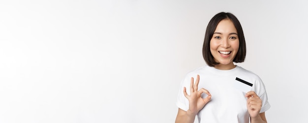 Ragazza coreana sorridente che mostra il segno giusto e che consiglia la carta di credito della banca dello spazio della copia che sta in ts