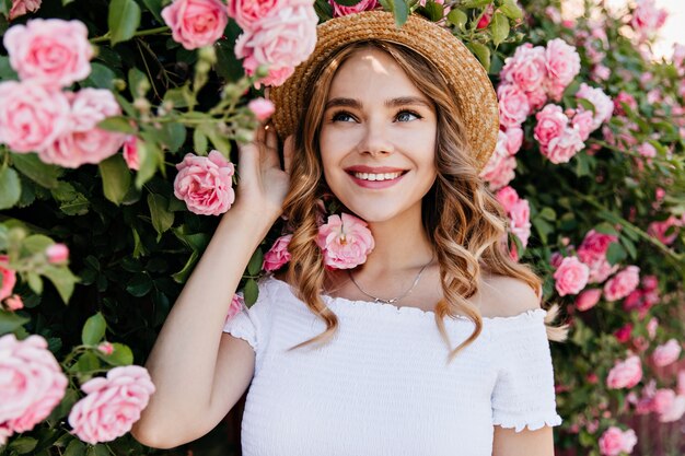 Ragazza dagli occhi azzurri accattivante in cappello estivo in posa in giardino. Ritratto all'aperto di donna riccia allegra che ride con le rose