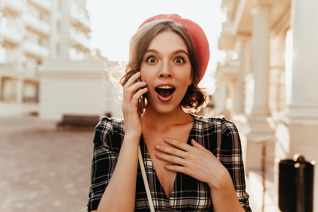 Ragazza dagli occhi scuri scioccata con un bel sorriso in piedi sulla strada. Donna riccia francese parlando al telefono in una giornata di sole.