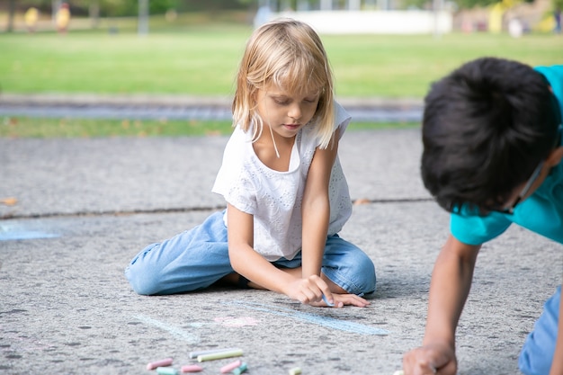 Ragazza dai capelli abbastanza bionda pensierosa che si siede e disegna con pezzi colorati di gessi. Vista frontale. Concetto di infanzia e creatività