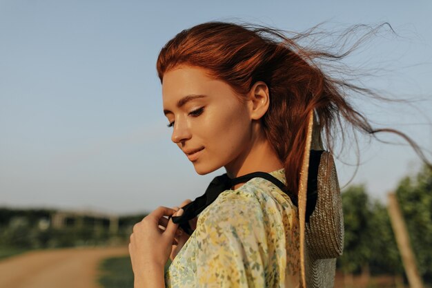 Ragazza dai capelli allo zenzero con graziose lentiggini, cappello di paglia e benda scura sul collo in abito giallo fresco che guarda in basso e posa all'aperto