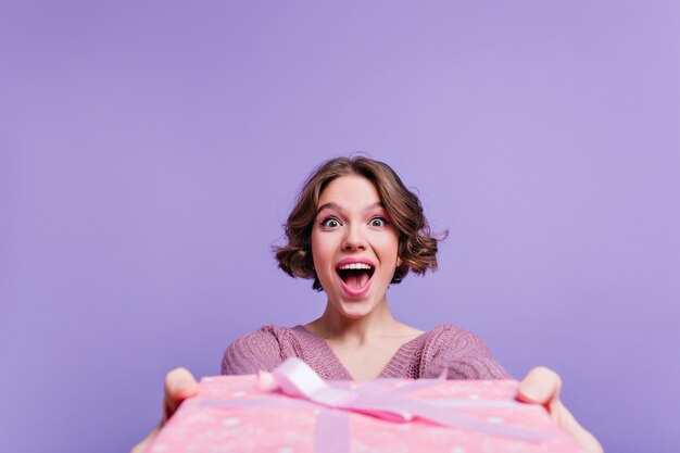 Ragazza dai capelli castani di risata isolata sulla parete viola con grande regalo di compleanno. Foto dell'interno della ragazza dai capelli corti carina che tiene il contenitore di regalo di Natale decorato con il nastro.