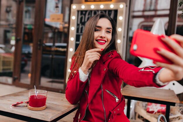 Ragazza dai capelli castani sognante utilizza lo smartphone per selfie sullo spazio urbano