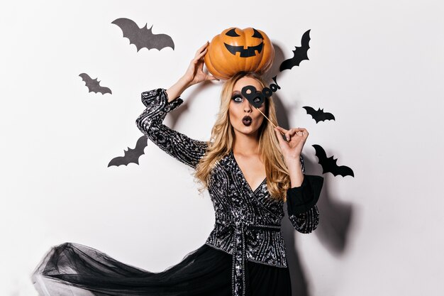 Ragazza dai capelli lunghi interessata che tiene la zucca arancione al servizio fotografico di Halloween. Foto interna di attraente signora bionda in costume da strega.