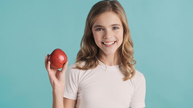 Ragazza dai capelli ondulati bionda allegra dell'adolescente che posa con la mela rossa che sorride sinceramente sulla macchina fotografica sopra fondo variopinto