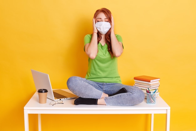 Ragazza dai capelli rossi in mascherina medica si siede con le gambe incrociate sul tavolo bianco, coprendosi le orecchie con i palmi e tiene gli occhi chiusi, indossa jeans e maglietta, circondato da lap top, caffè, penne