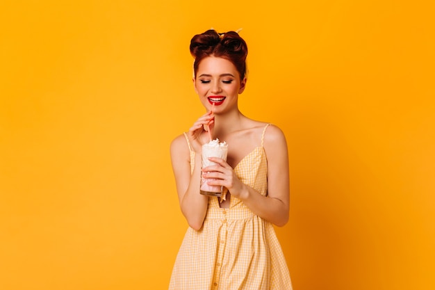 Ragazza dai capelli rossi positiva che beve milkshake. Studio shot di blithesome pinup lady isolato su spazio giallo.