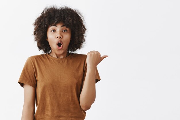 Ragazza dalla pelle scura impressionata e sbalordita con un'acconciatura afro in marrone alla moda con le labbra aperte pieghevoli che puntano a destra con il pollice che descrive la scena fantastica che ha visto in quella direzione