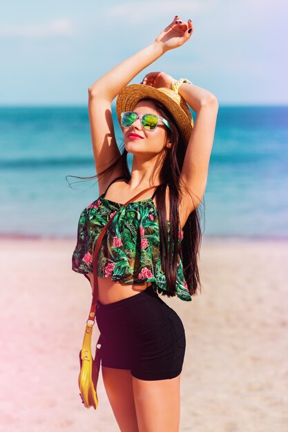 Ragazza dalla vestibilità perfetta in occhiali da sole alla moda con corpo abbronzato in posa sulla spiaggia tropicale con top colorato brillante, pantaloncini alti ed eleganti accessori swag.