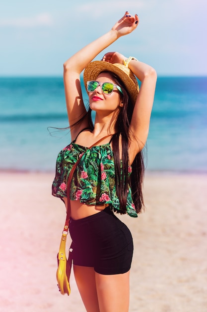 Ragazza dalla vestibilità perfetta in occhiali da sole alla moda con corpo abbronzato in posa sulla spiaggia tropicale con top colorato brillante, pantaloncini alti ed eleganti accessori swag.