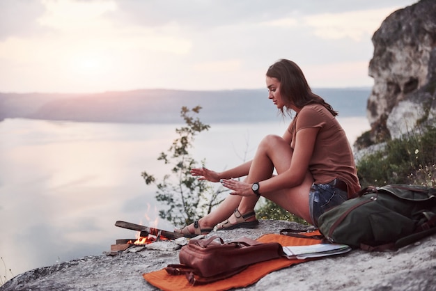 Ragazza dei pantaloni a vita bassa con lo zaino che gode del tramonto sul picco della montagna della roccia.