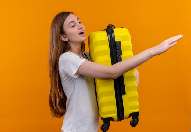 Ragazza del giovane viaggiatore che tiene la valigia che allunga la mano e che esamina il lato destro sulla parete arancione isolata con lo spazio della copia