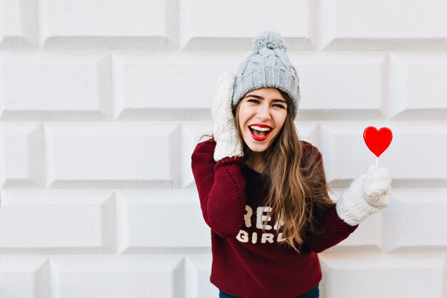 Ragazza del ritratto con capelli lunghi in cappello lavorato a maglia e maglione marsala con lecca-lecca cuore rosso sul muro grigio. Indossa guanti bianchi caldi, ridendo.