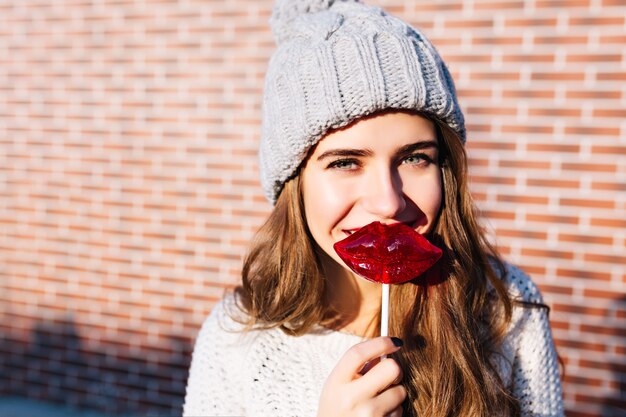 Ragazza del ritratto del primo piano con capelli lunghi in cappello lavorato a maglia con le labbra rosse della lecca-lecca sulla parete esterna. Lei sta sorridendo .