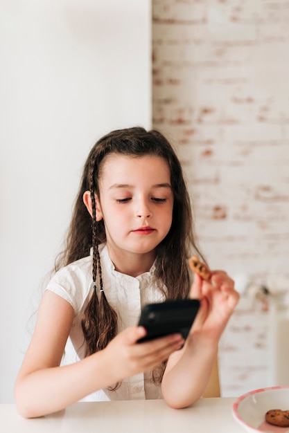 Ragazza del tiro medio con il telefono che mangia i biscotti