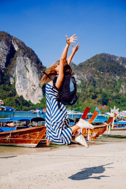 Ragazza del viaggiatore con zaino e sacco a pelo abbastanza tan che posa all'isola tropicale calda di Phi Phi, vista stupefacente sulle barche e sulle montagne locali