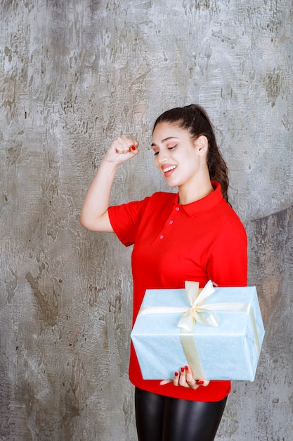 Ragazza dell'adolescente che tiene una scatola regalo blu avvolta con un nastro bianco e che mostra il segno della mano di godimento.