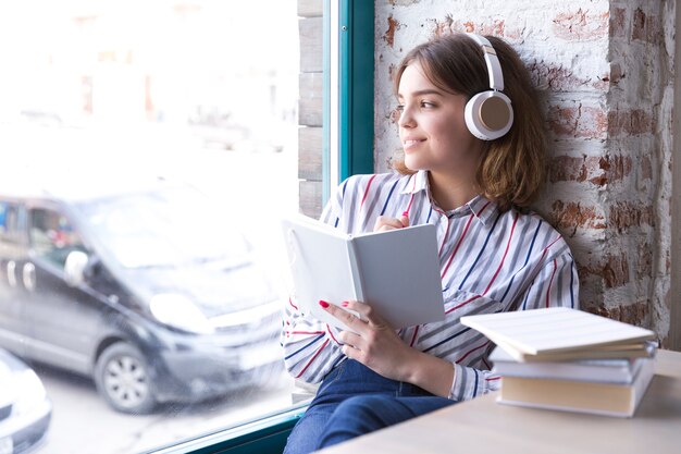 Ragazza dell&#39;adolescente in cuffie che si siedono con il libro aperto che osserva fuori finestra