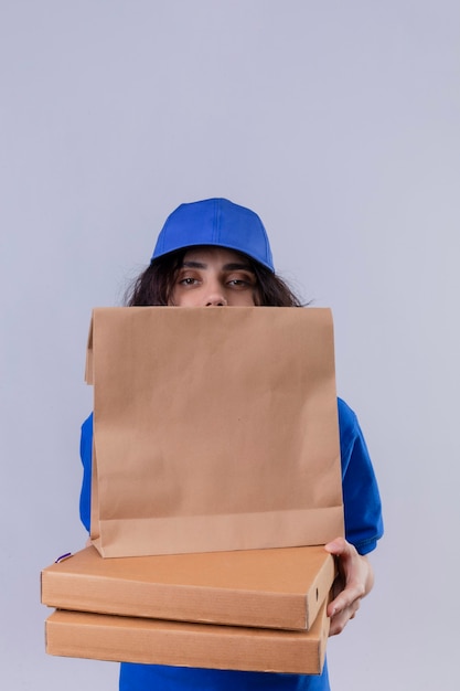 Ragazza delle consegne in uniforme blu che si nasconde dietro le scatole della pizza e il pacchetto di carta in piedi