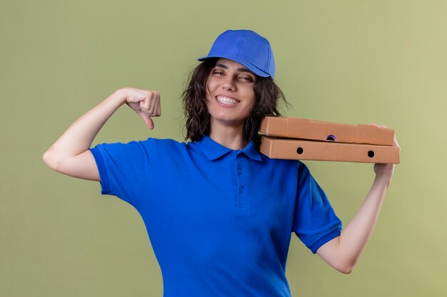 Ragazza delle consegne in uniforme blu che tiene le scatole per pizza che mostrano il bicipite sorridente allegramente in piedi sopra lo spazio verde isolato