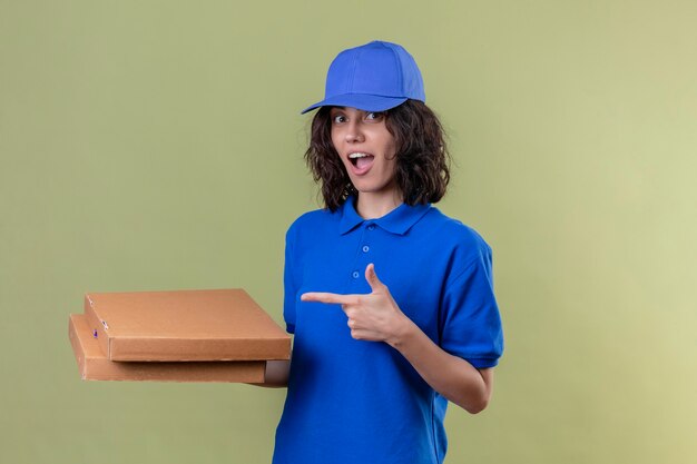 Ragazza delle consegne in uniforme blu che tiene le scatole per pizza e punta il dito indice a loro sorridendo con la faccia felice in piedi sopra lo spazio verde