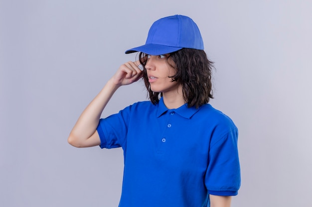 Ragazza delle consegne in uniforme blu e cappuccio che guarda lontano con espressione confusa in piedi sul bianco isolato