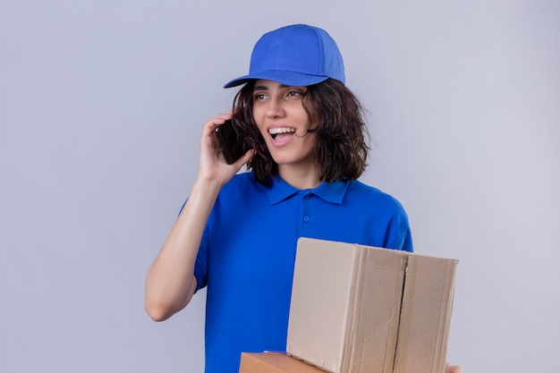 Ragazza delle consegne in uniforme blu e cappuccio che tiene le scatole della pizza e il pacchetto della scatola e parla al telefono cellulare con la faccia felice che sta sul bianco