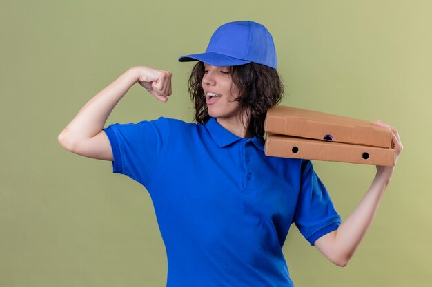 Ragazza delle consegne in uniforme blu e cappuccio che tiene le scatole per pizza che mostrano il bicipite che sorride allegramente in piedi sul verde isolato