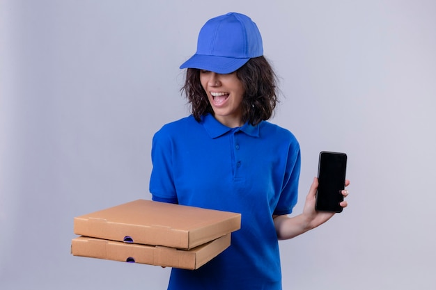 Ragazza delle consegne in uniforme blu e cappuccio che tiene le scatole per pizza sorridendo allegramente mostrando il telefono cellulare in piedi