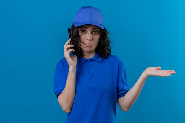 Ragazza delle consegne in uniforme blu e cappuccio parlando al telefono cellulare all'oscuro e confuso con le braccia aperte, nessun concetto di idea in piedi sul blu isolato