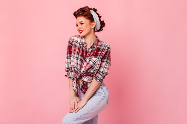 Ragazza dello zenzero Debonair in camicia a scacchi ballando con un sorriso. Studio shot di pinup donna in blue jeans isolato su uno spazio rosa.