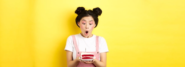 Ragazza di compleanno asiatica sveglia con trucco luminoso che soffia candela sulla torta che fa desiderio che sta sul sedere giallo