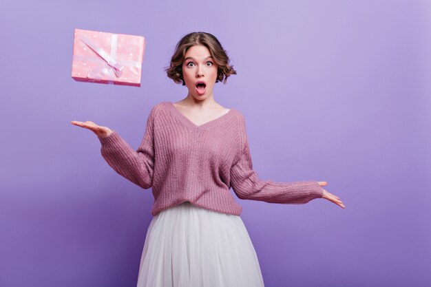 Ragazza di compleanno divertente in maglione che esprime emozioni sorprese sulla parete viola. Emotiva signora dai capelli corti in gonna bianca in posa con la presente casella a casa.