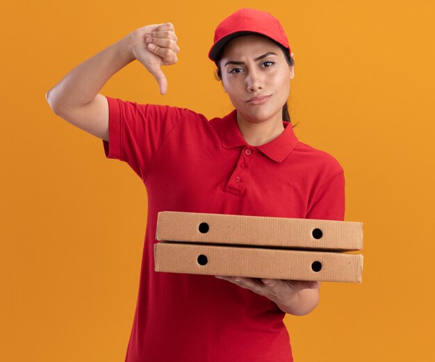 Ragazza di consegna giovane dispiaciuta che indossa l'uniforme e il cappuccio che tengono le scatole per pizza che mostrano il pollice verso il basso isolato sulla parete arancione